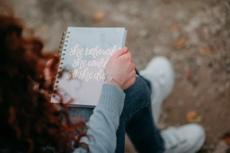 A woman holds a notebook with an inspirational quote, sitting outdoors.